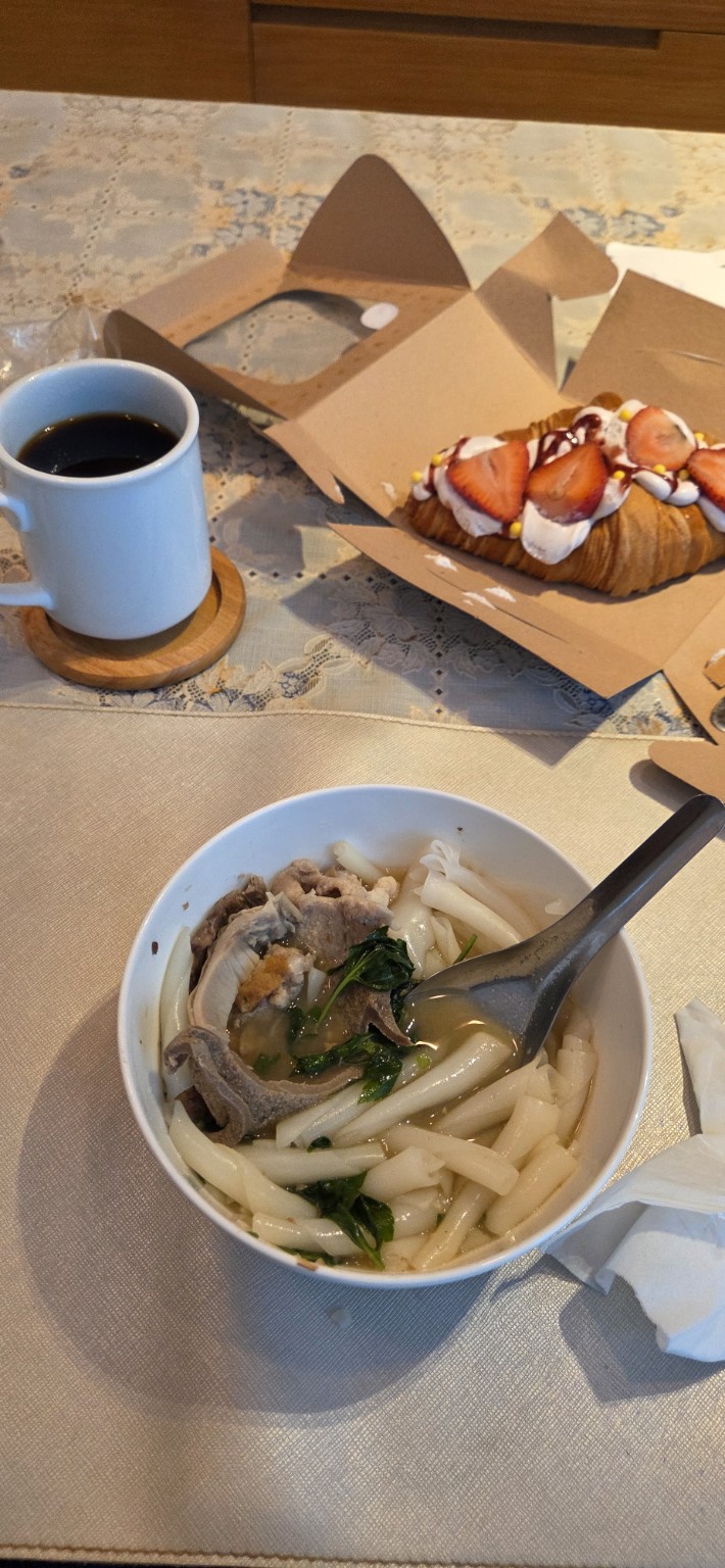 Bowl of wide rice noodles with pork beside coffee and a strawberry pastry